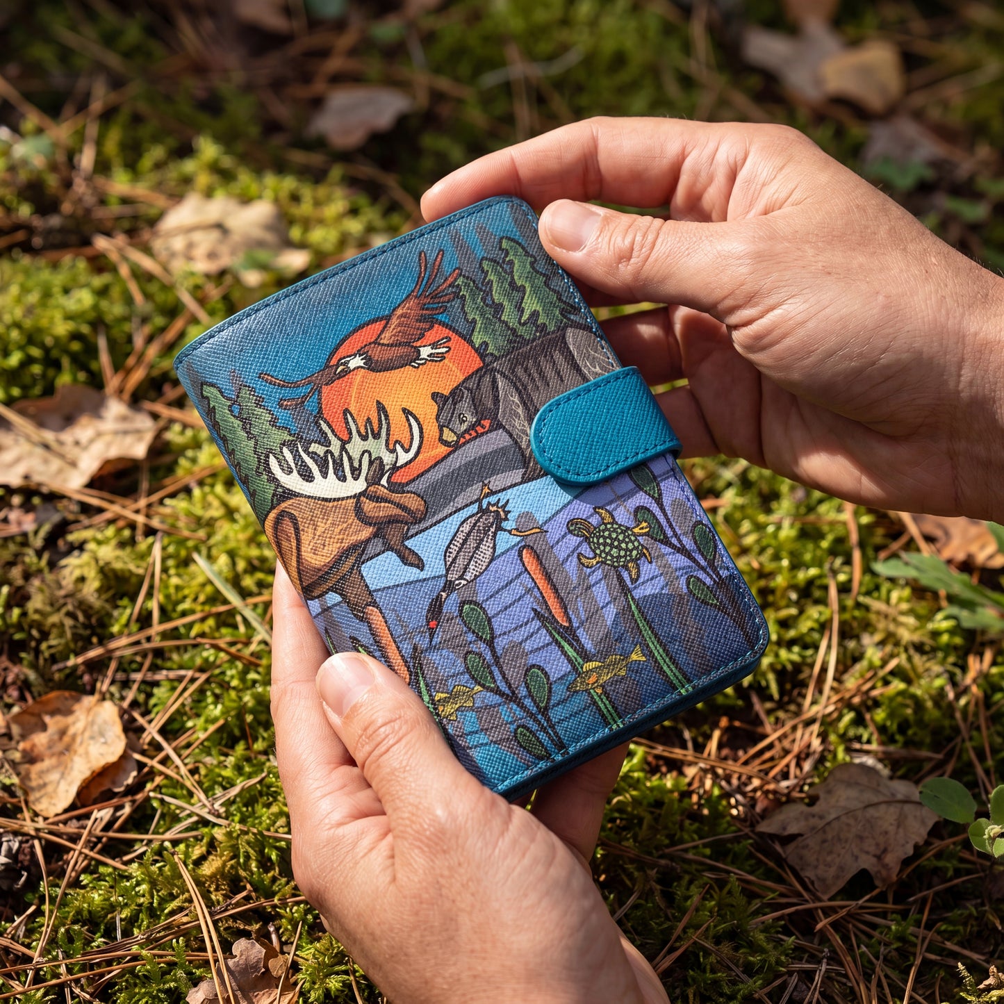 Hand holding a blue wallet with nature-themed design on a mossy ground background