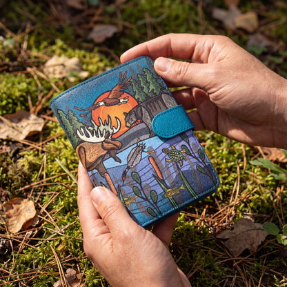 Hand holding a blue wallet with nature-themed design on a mossy ground background
