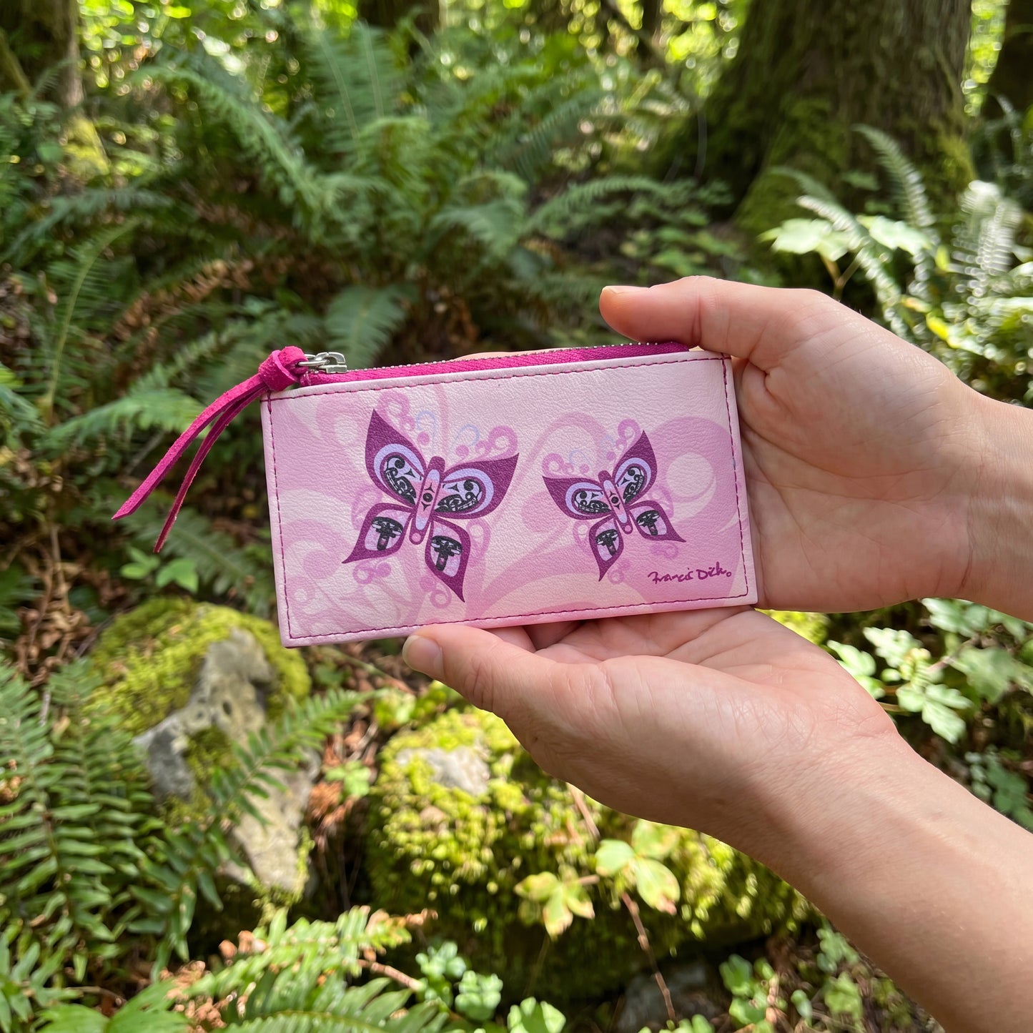 Hand holding a pink butterfly-themed pouch in front of a forest background