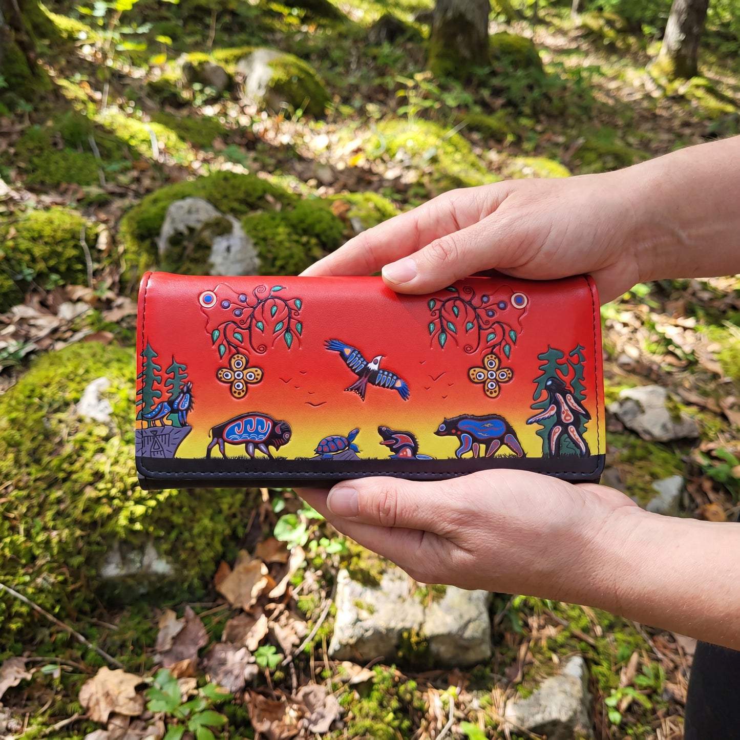 Hand holding a red wallet with nature-themed designs against a forest background