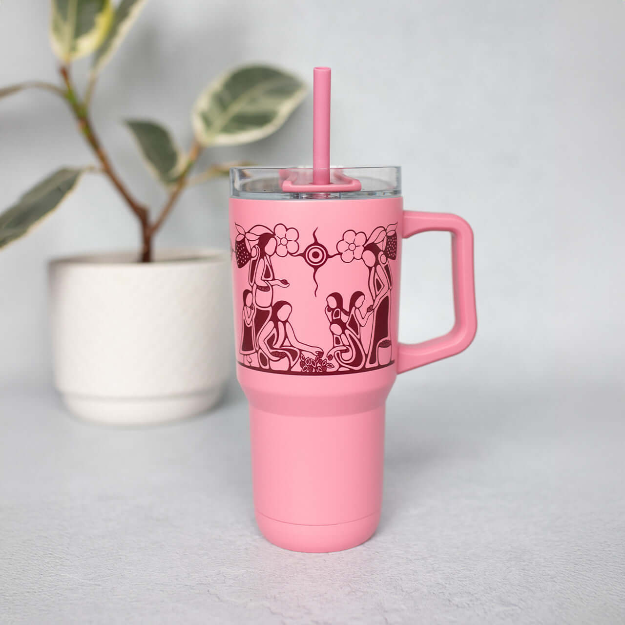 Heart Berry Gatherers insulated tumbler with pink Indigenous design on the table near plant