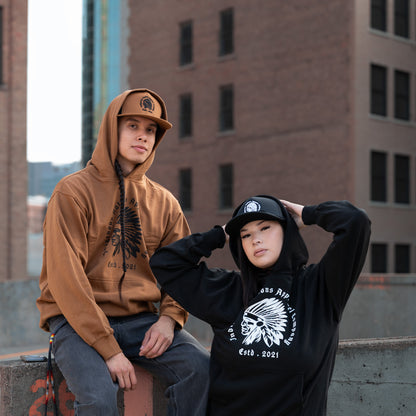Two models posing in urban streetwear on a rooftop with brick buildings in the background. One wears a brown hoodie and cap, and the other wears a black hoodie with a white graphic and a black cap.