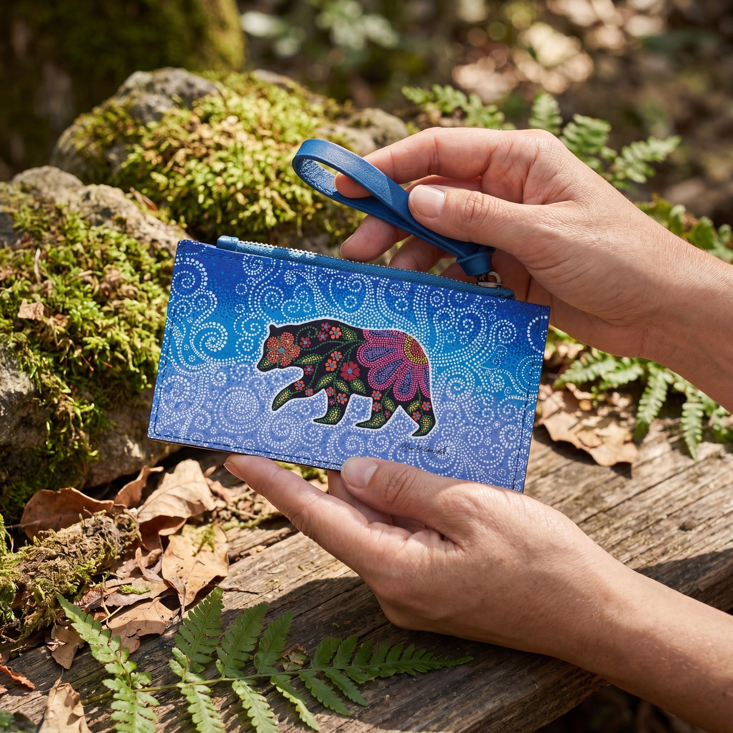 Person holding a blue pouch with a colorful bear design in a natural setting.