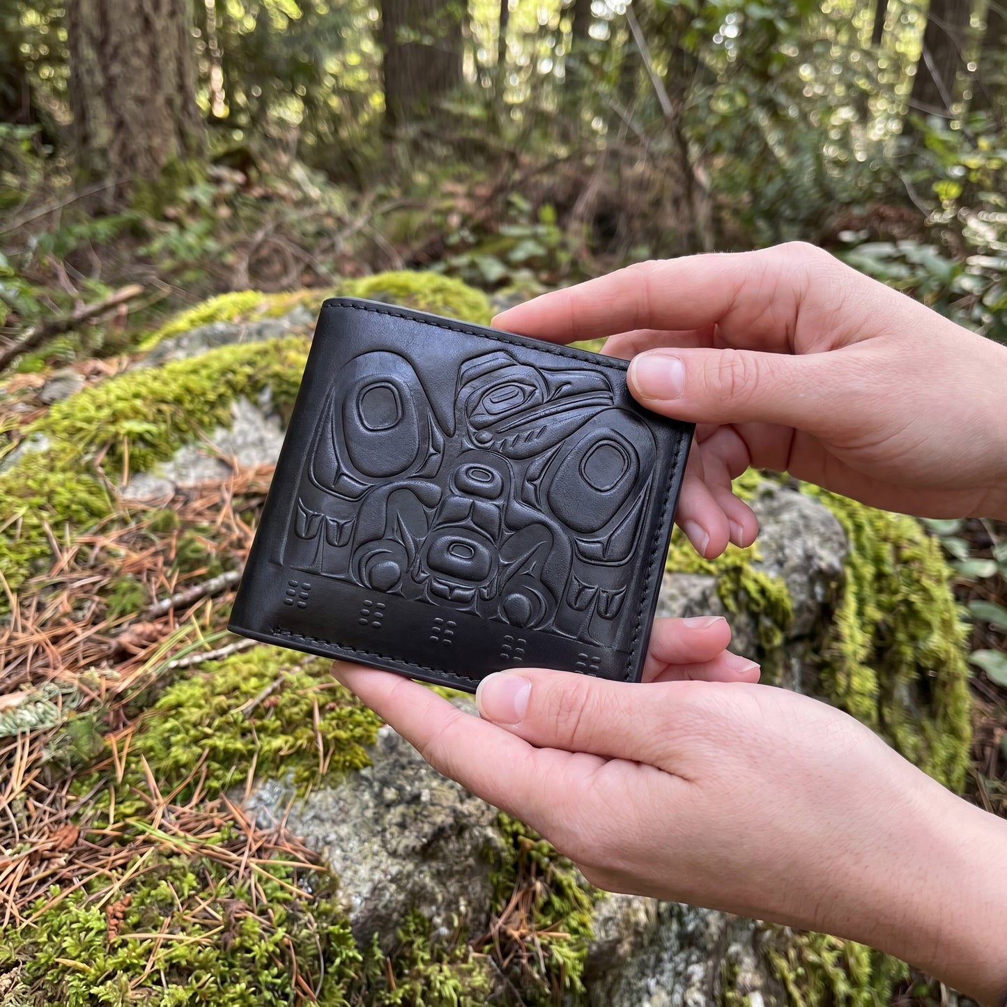 Person holding a black leather wallet with tribal design in a forest setting