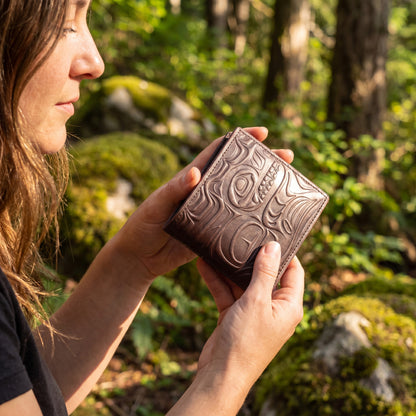 Person holding a brown leather wallet with intricate designs in a forest setting