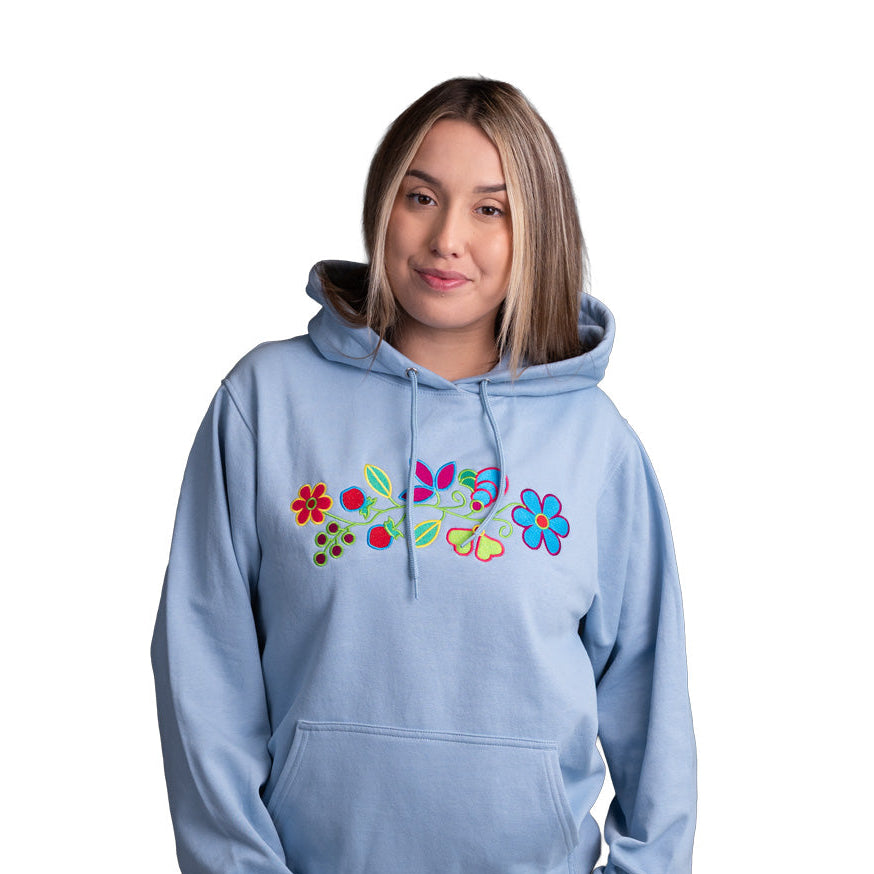 A woman modeling a light blue pullover hoodie with a colorful embroidered floral design on the chest, set against a plain white background.